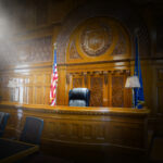 Wood-paneled courtroom with a raised judge's bench, American flag, and a blue flag, softly lit with atmospheric light.