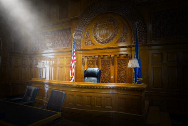 Wood-paneled courtroom with a raised judge's bench, american flag, and a blue flag, softly lit with atmospheric light.