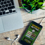 Laptop keyboard visible on a wooden desk next to a smartphone displaying a sports betting app.