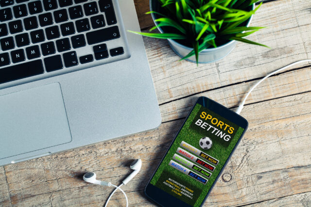 Laptop keyboard visible on a wooden desk next to a smartphone displaying a sports betting app.