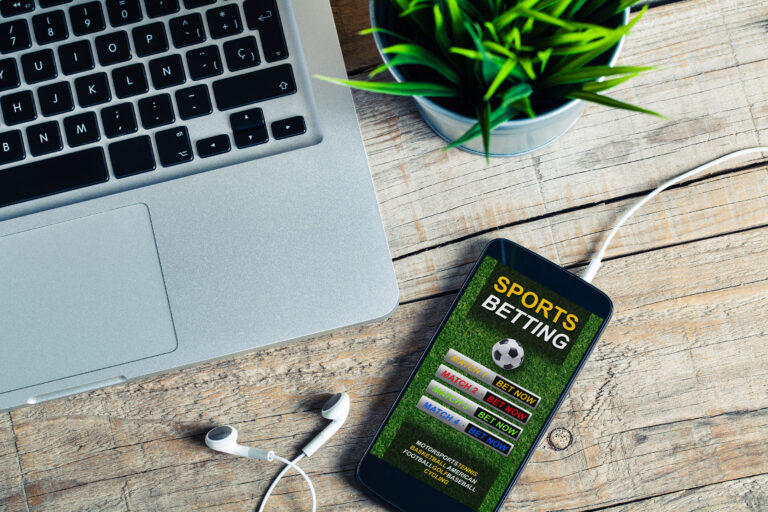 Laptop keyboard visible on a wooden desk next to a smartphone displaying a sports betting app.