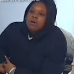 Woman in a black hoodie sitting on a sofa, looking surprised off-camera.