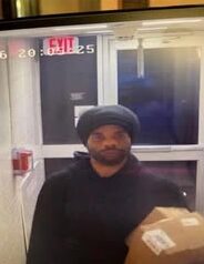 Man in a dark hoodie wearing a cap stands inside a doorway, holding a cardboard box