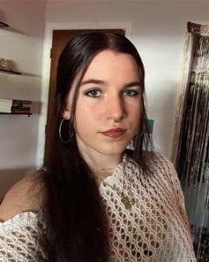 Portrait of a young woman with long dark hair and hoop earrings, wearing a light crochet top, posing indoors.
