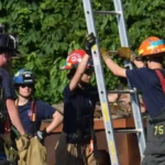 Firefighters in turnout gear practice a rescue drill, stabilizing a tall ladder against a building.