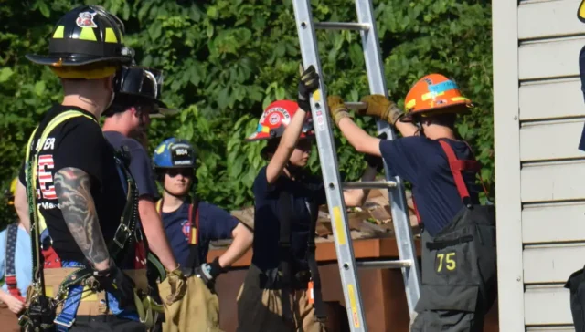 Firefighters in turnout gear practice a rescue drill, stabilizing a tall ladder against a building.