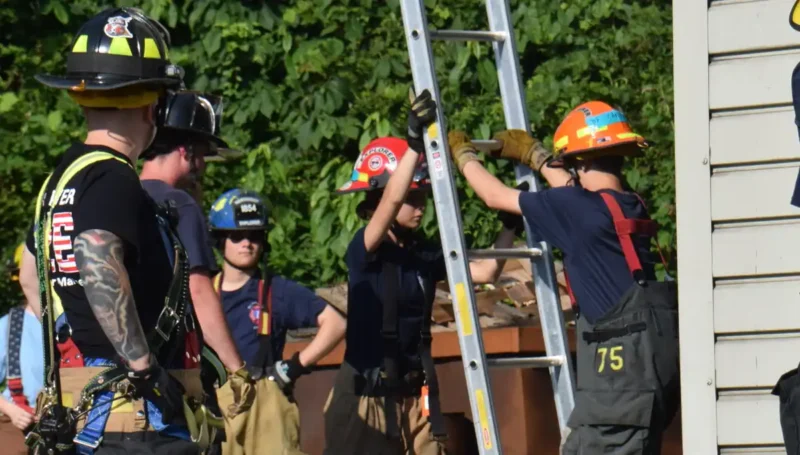 Firefighters in turnout gear practice a rescue drill, stabilizing a tall ladder against a building.